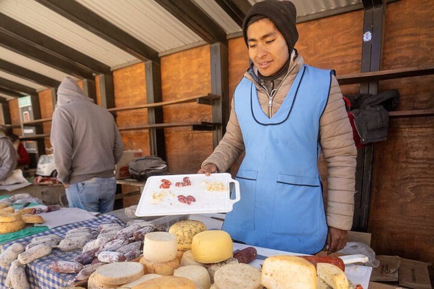 A person at a market stall with various cheeses and cured meats. 3