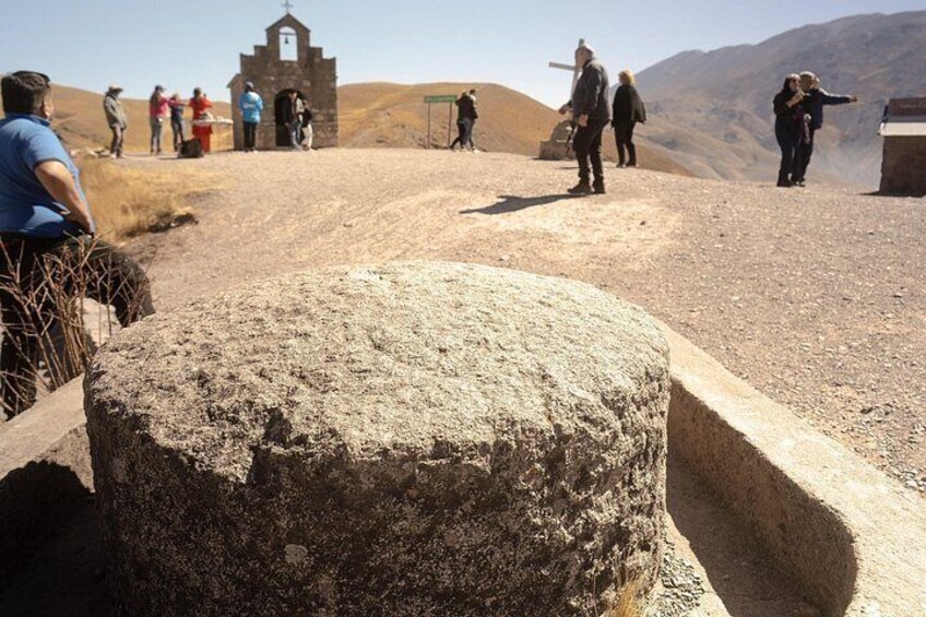 Un paisaje rocoso con un gran bloque de piedra en primer plano, un pequeño edificio con una cruz y personas caminando al fondo. 2