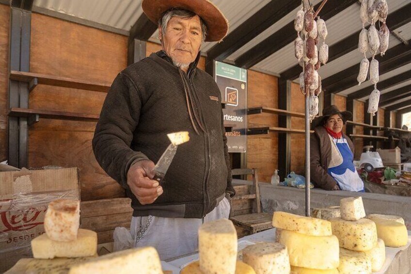 Una persona cortando queso en un puesto de mercado, con varios quesos y salchichas colgando al fondo. 5