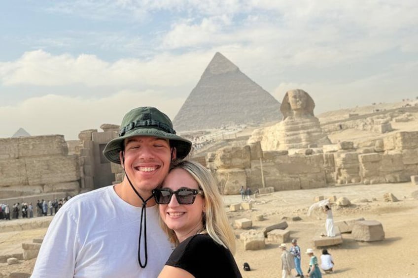 Een persoon in een wit shirt en groene hoed staat voor de Piramide van Gizeh, met de Sfinx op de achtergrond. 1