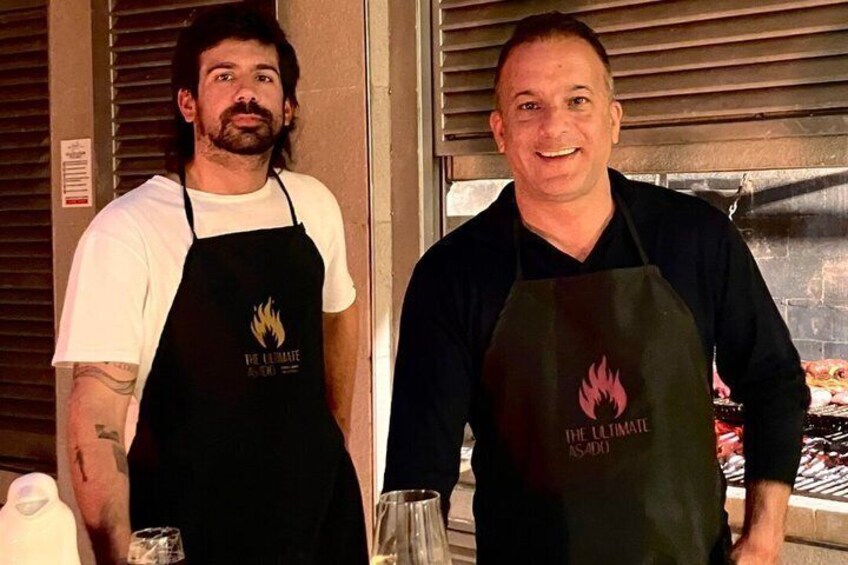 Two people wearing aprons with a flame logo, standing in front of a grill. 2
