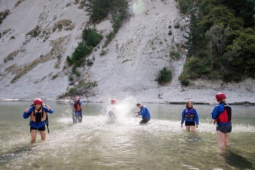 Mensen in blauwe jacks en rode helmen waden door ondiep water nabij een rotsachtige heuvel. 2