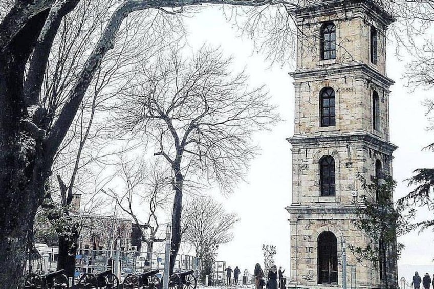 A historic stone tower with multiple windows and a clock, surrounded by leafless trees and a crowd of people. 4