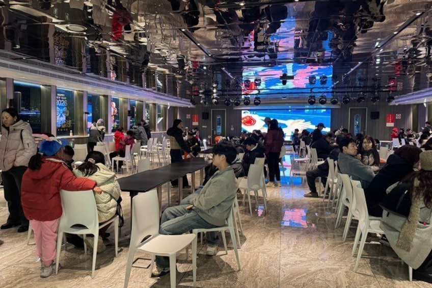 A crowded indoor space with people seated at tables, a large screen displaying an image, and a ceiling with hanging lights. 3
