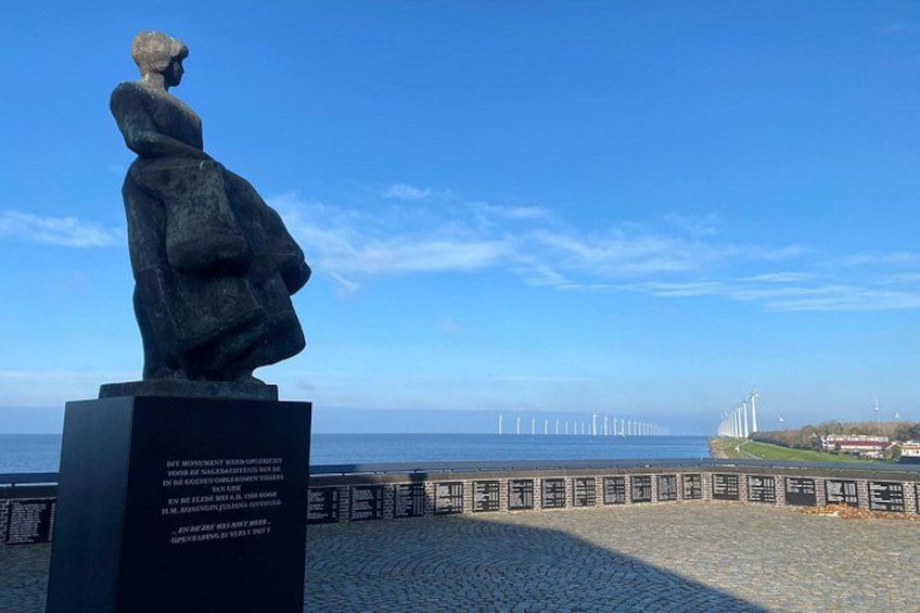 Una estatua de una mujer sobre un pedestal, con vista al mar, aerogeneradores a lo lejos y un camino pavimentado. 5