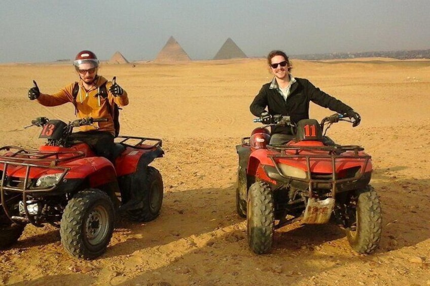 Two people on ATVs in a desert with pyramids in the background. 2