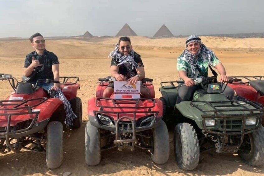 Three people on ATVs in a desert with pyramids in the background. 4