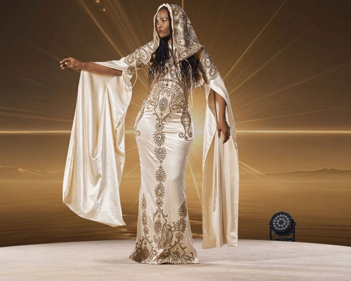A person in a detailed white garment with gold embroidery, standing in a desert-like setting with a chair and a light fixture in the background. 3