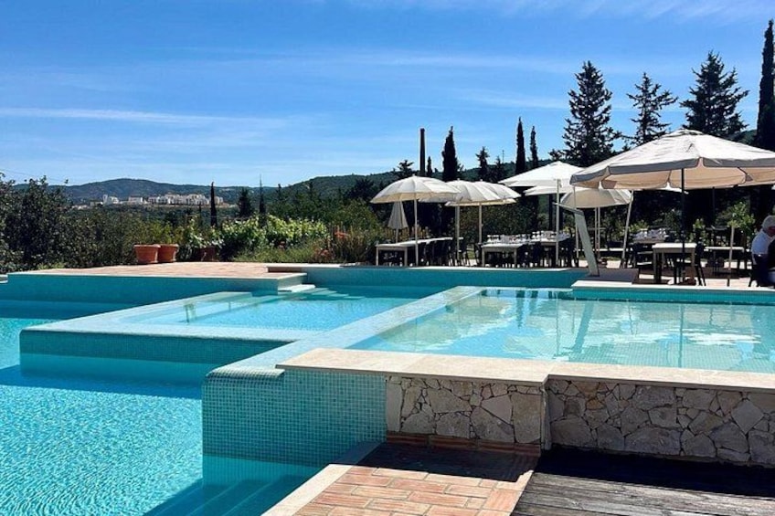 Una piscina con bordi piastrellati, circondata da sedute all'aperto e ombrelloni, con vista su alberi e colline. 3
