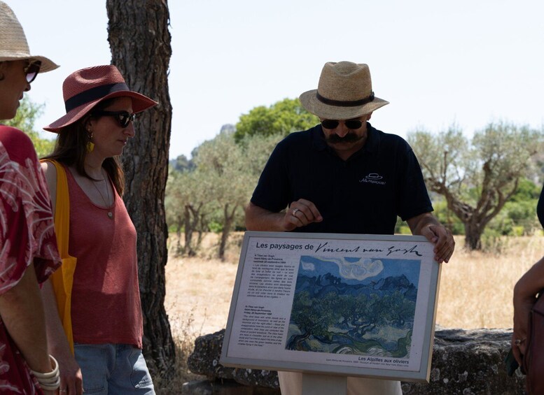 Una persona sostiene un cartel con una pintura de paisaje y texto, de pie al aire libre con árboles y un cielo despejado de fondo. 4