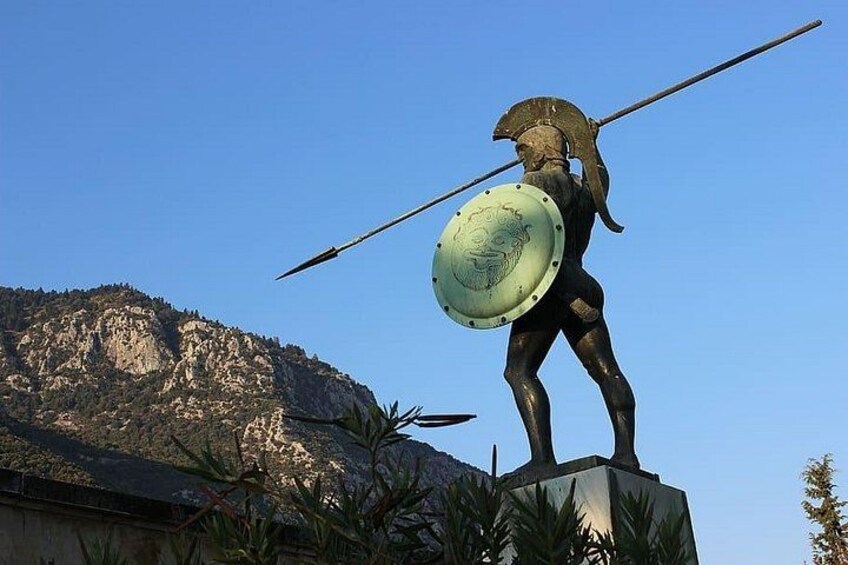 A bronze statue of a warrior holding a spear and shield, standing on a pedestal with a mountainous backdrop. 1