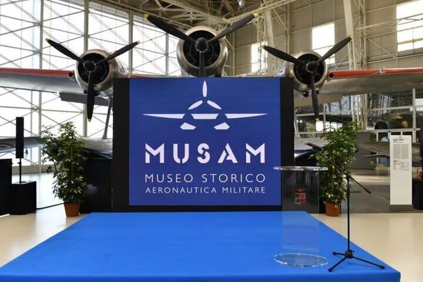 A display table with a blue cloth, a sign reading "MUSAM Museo Storico Aeronautica Militare", and three aircraft engines mounted above. 1