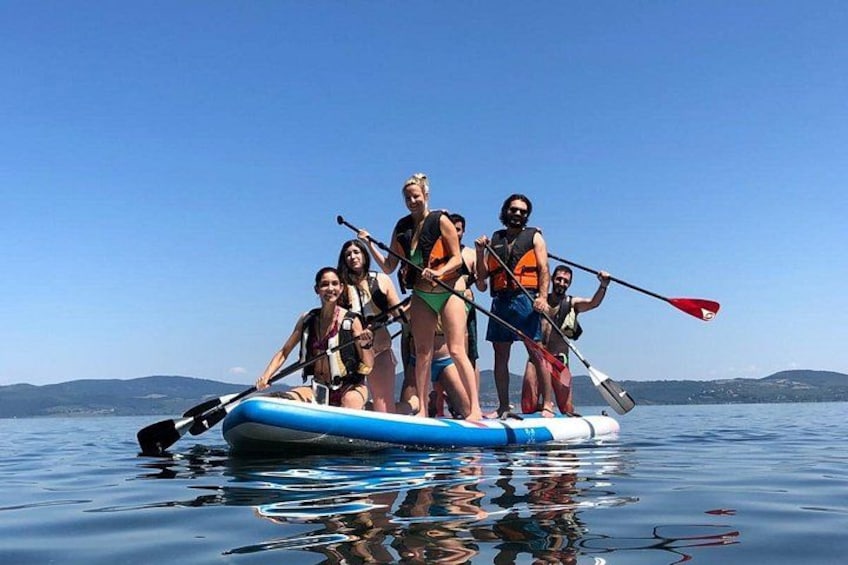 A group of people on a blue raft paddleboarding on calm water with a clear sky and distant hills. 1