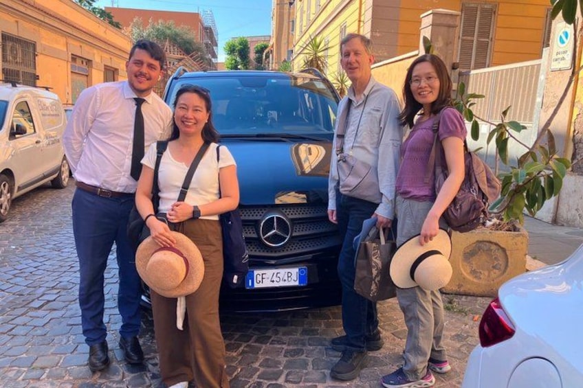 A group of people standing in front of a blue car with a license plate, a cobblestone street, and buildings in the background. 2