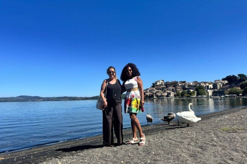 Two people standing on a pebbled shore by a body of water with a swan and ducks nearby. 3
