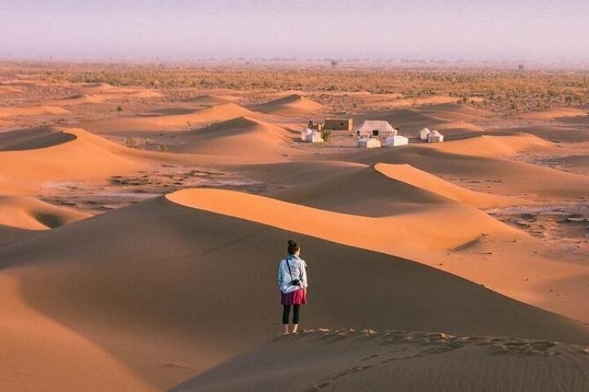 A person standing on a sand dune with a desert landscape and small buildings in the distance. 1