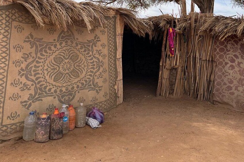 A traditional hut with a patterned wall and thatched roof, a dirt ground, and several plastic containers outside. 4
