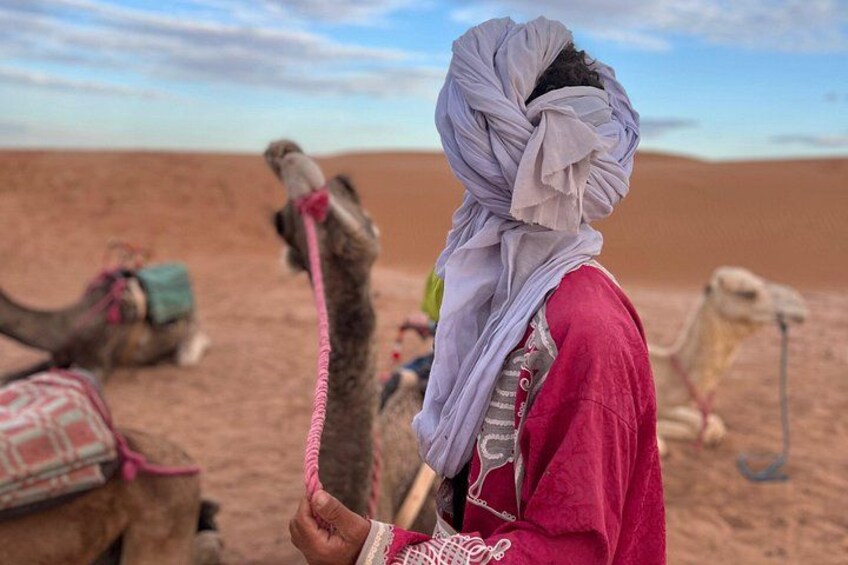 A person in traditional attire with a camel in a desert landscape. 2