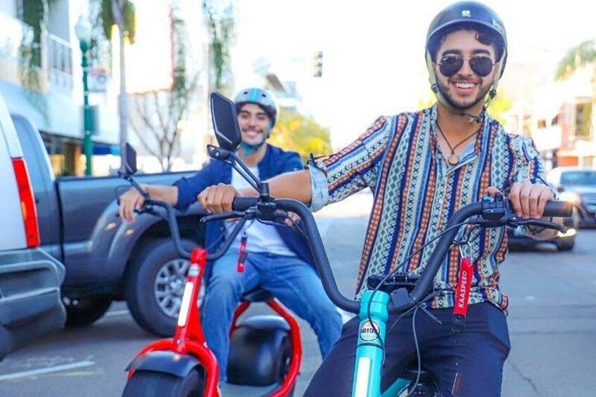 Two people riding electric scooters on a street with cars and buildings in the background. 2