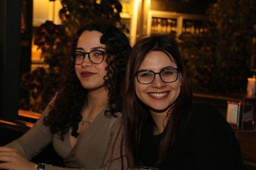 Two people sitting at a table in a dimly lit indoor setting with plants and a bar in the background. 3