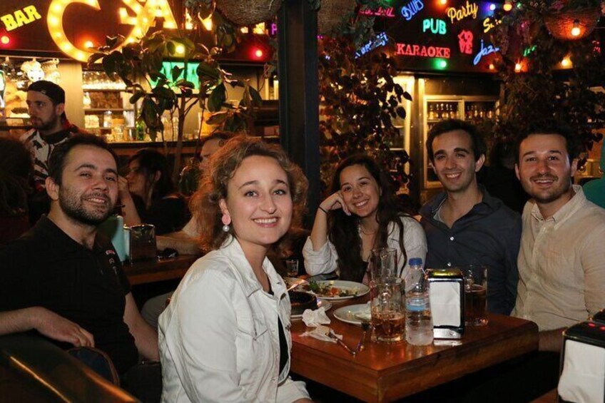 Un groupe de personnes est assis à une table dans un restaurant qui dispose d’un bar et où l’on peut voir, au fond, une enseigne indiquant qu’il y a un karaoké. 4