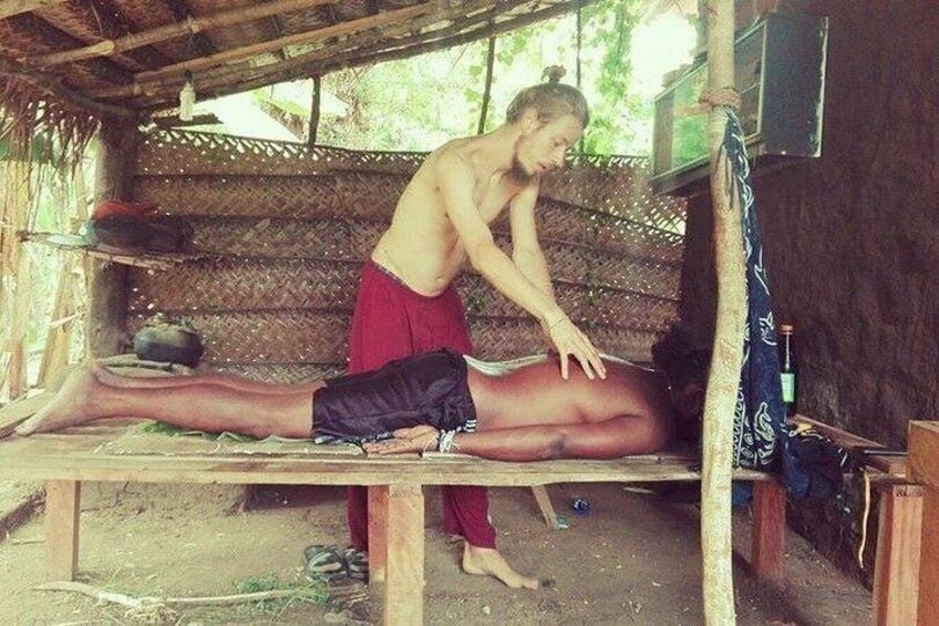 A person lying on a wooden bench outdoors, another person standing beside them, and a hut-like structure in the background. 1