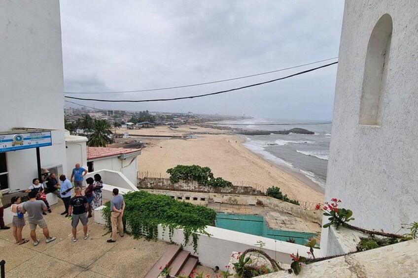 A beach with people standing on a balcony overlooking the ocean, a pool, and a building with a sign. 2