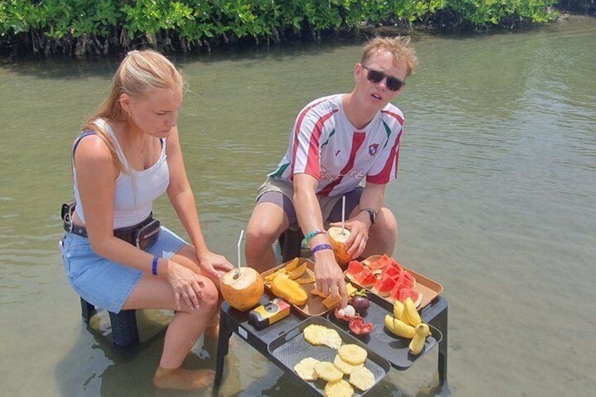 To personer sidder ved et bord med mad og drikke i et vandområde omgivet af grønt. 5