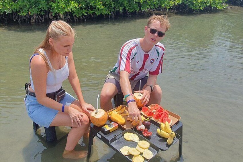 Two people sitting by a table with food and drinks in a body of water surrounded by greenery. 5
