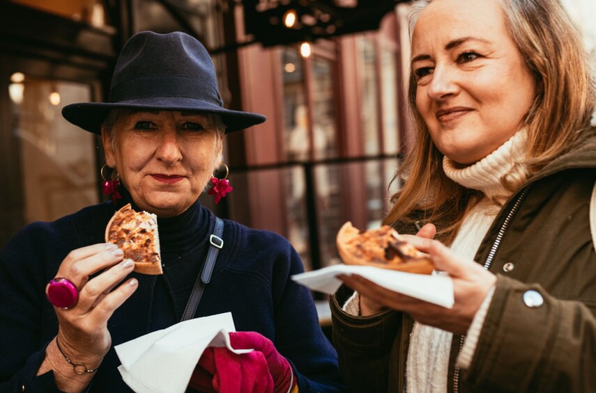 Two people holding slices of pizza, one wearing a hat and the other in a jacket, in an urban setting with buildings in the background. 1