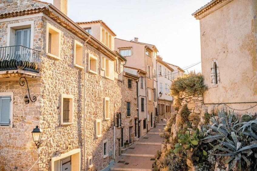 A narrow stone street flanked by buildings with shutters, a lamp post, and a plant. 5