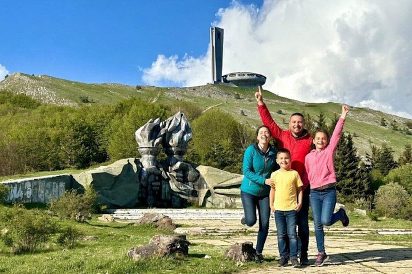 Una famiglia posa davanti a una grande scultura e un edificio alto in un paesaggio collinare. 1