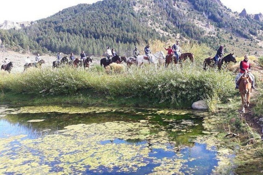 Un gruppo di persone cavalca lungo un argine fluviale, con montagne sullo sfondo. 5