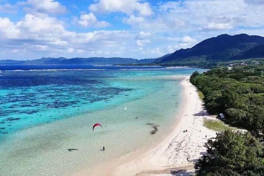 Een strand met helder turquoise water, een persoon paraglide en een bergachtig landschap op de achtergrond. 1
