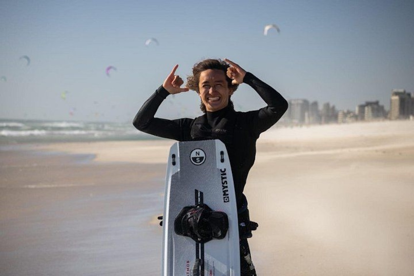 Een persoon in een neopreenpak houdt een surfplank vast, met het merk 'DYSTIC' erop, op een strand met een stadsachtergrond. 3
