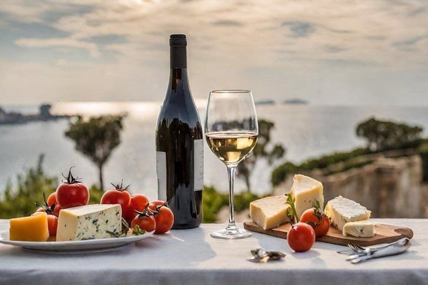 A table with cheese, tomatoes, a bottle of wine, and a glass of wine with a scenic view of the sea and cliffs in the background. 3
