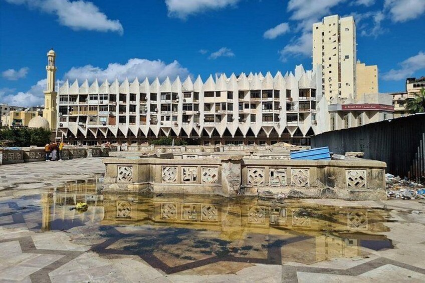 A building with a unique geometric facade, a reflective water feature, and a tall minaret in the background. 2