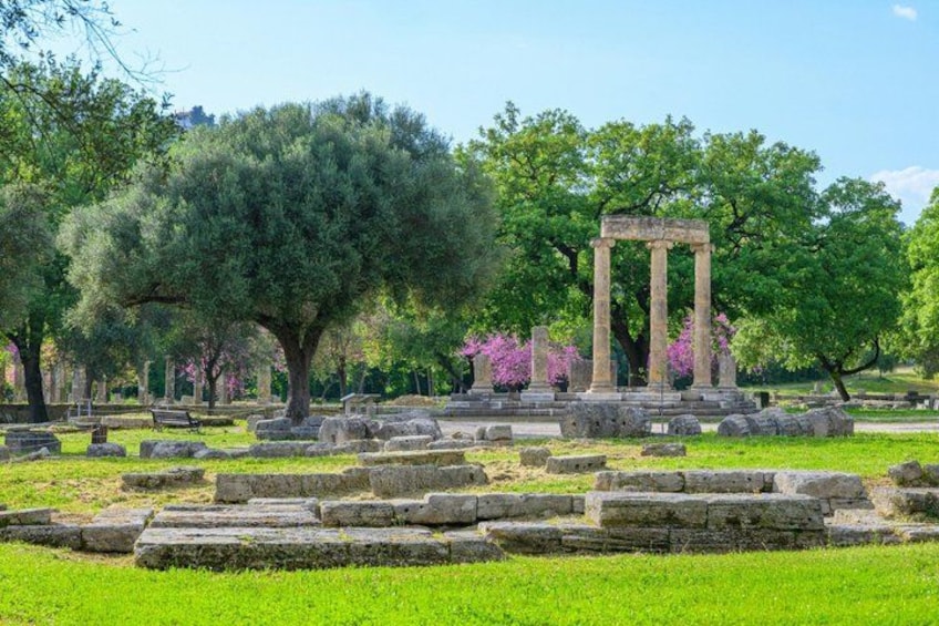 Ancient ruins with columns and stone blocks, surrounded by green grass and trees. 2
