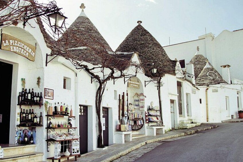 Una strada con edifici bianchi, un negozio con bottiglie di vino e un cartello che recita 'LA PIZZICHERIA ENOTECA'. 1