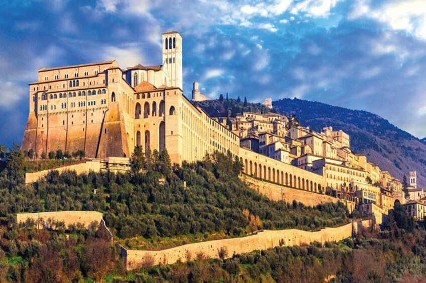A historic building with a prominent tower, situated on a hillside with a dense forest in the foreground. 1