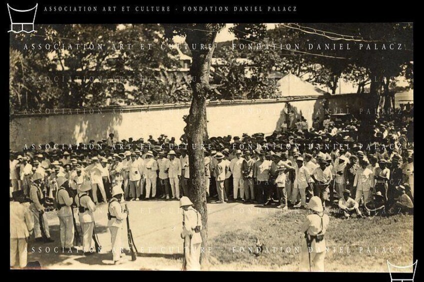 A group of people gathered outdoors, some standing and some sitting, with trees and a building in the background. 2
