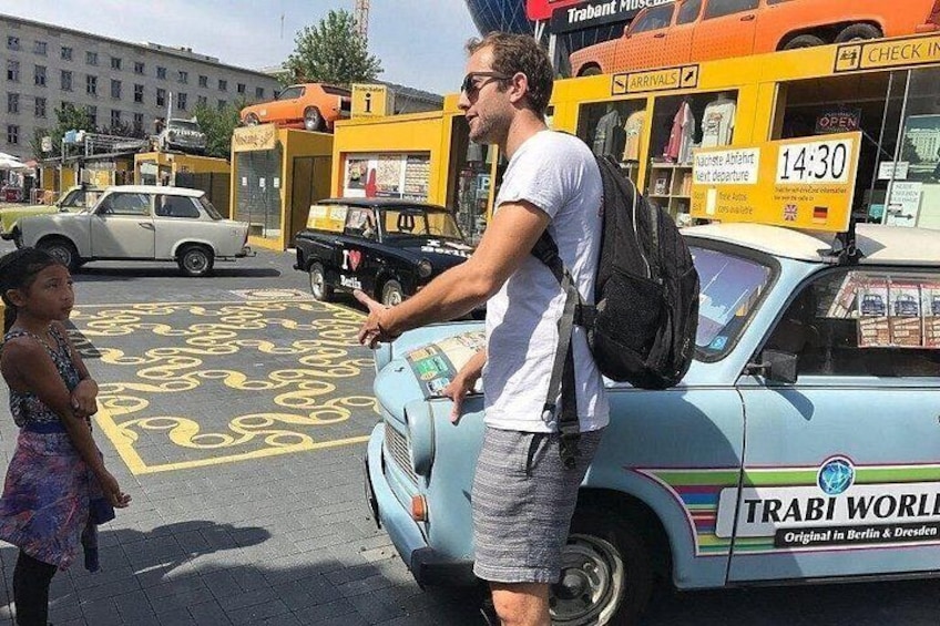 A vintage blue taxi with 'TRABI WORLD' written on it, parked on a street with yellow markings. 1