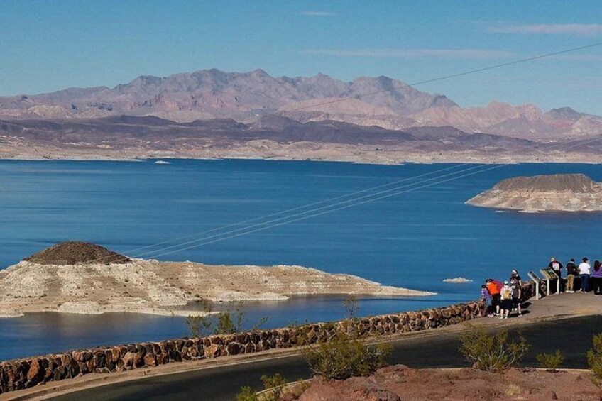 Un lago con montañas al fondo, una plataforma de observación con gente y una costa rocosa. 5