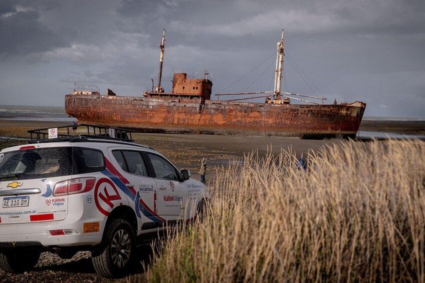 En hvid bil med racerklistermærker parkeret foran et rustent skib på stranden. 2