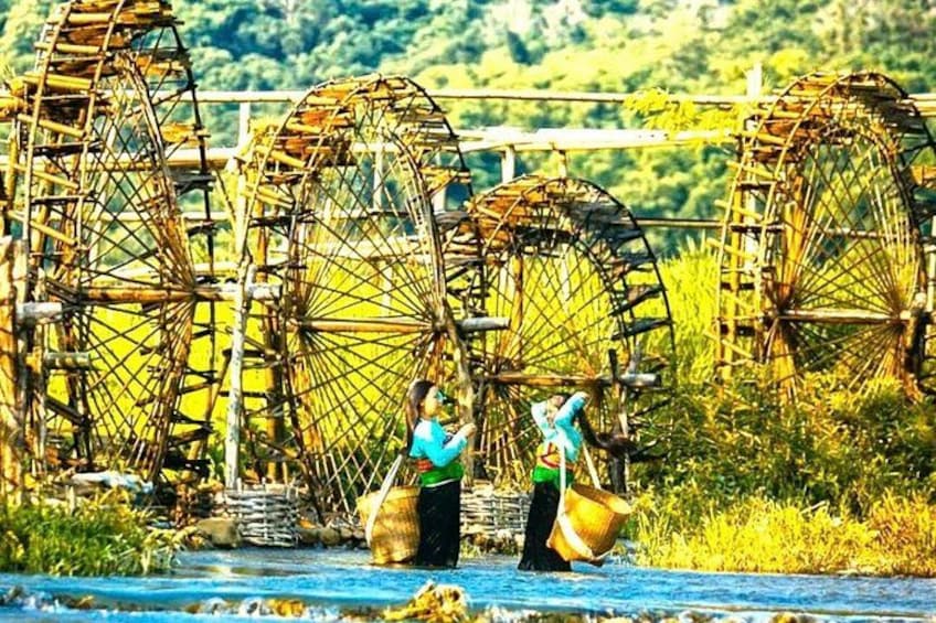 A river with large water wheels, a person carrying a basket, and another person with a bag. 1