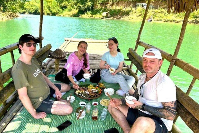 A group of people dining on a boat with a bamboo structure, surrounded by a calm river. 4