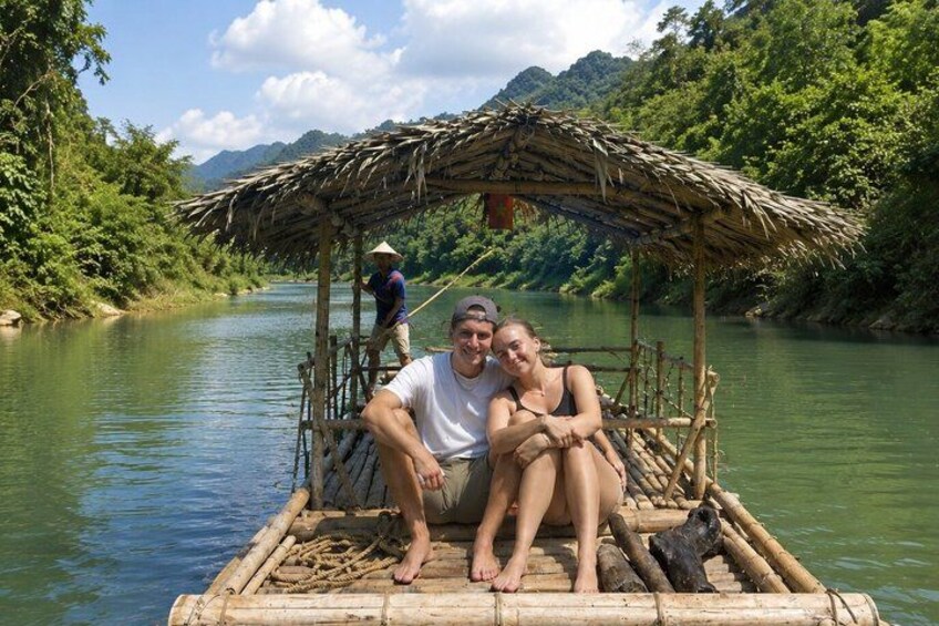 A couple on a bamboo raft with a thatched roof, a person standing on the riverbank, and lush greenery along the river. 4