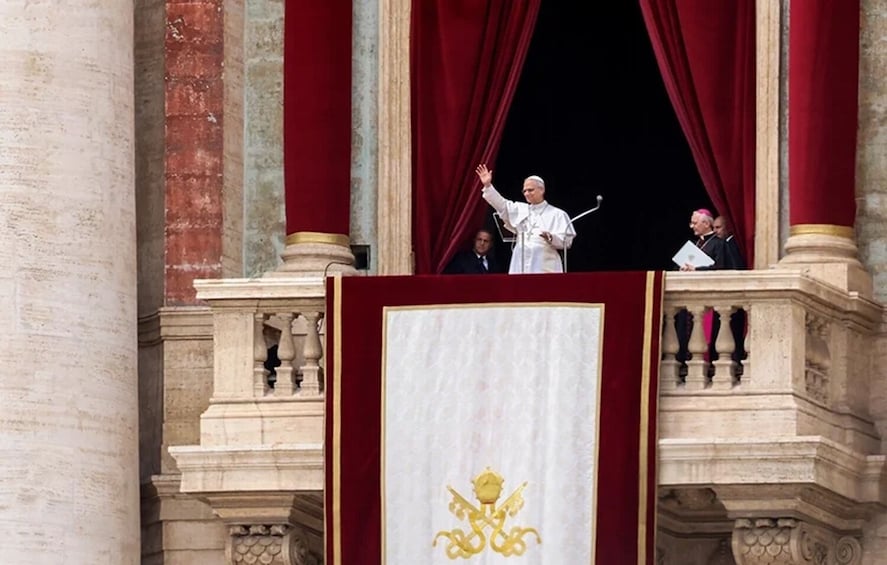 A person standing on a balcony with a red curtain backdrop, a white podium with a golden emblem, and architectural columns. 1