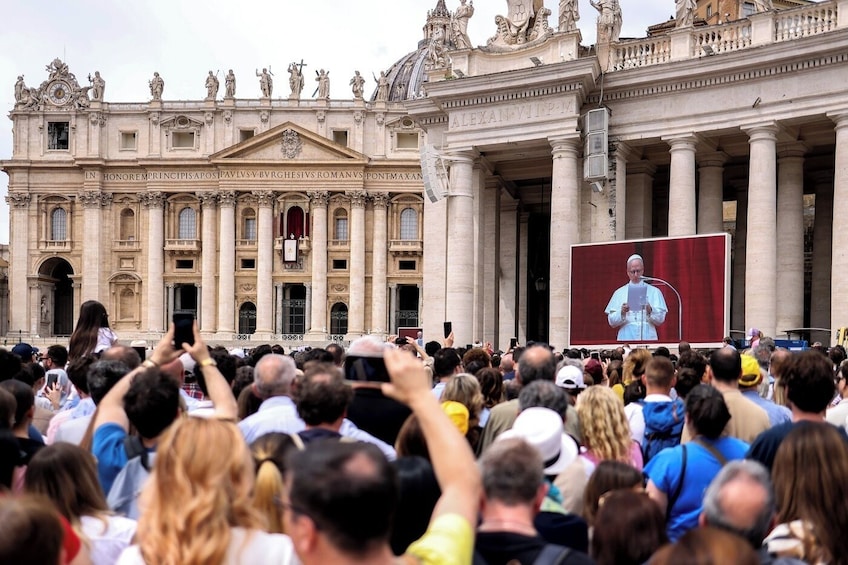 A crowd in front of a grand building with columns and statues, a large screen displaying a person speaking, and a crowd holding up phones. 5