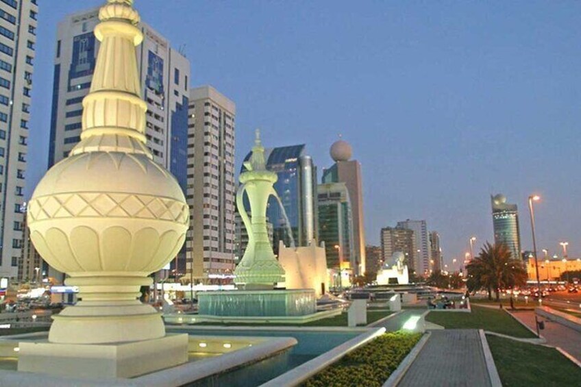 A cityscape with modern buildings, a large illuminated fountain, and a decorative structure resembling a vase. 3